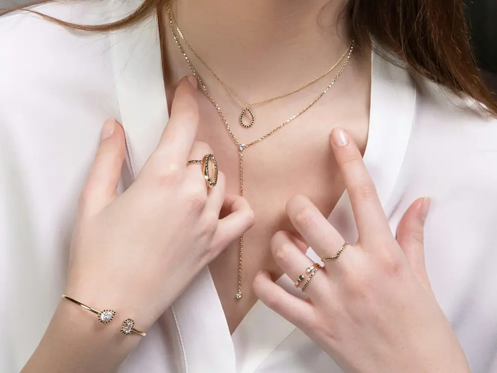 Mulher de camisa branca com colares finos em camadas, anéis e bracelete dourados